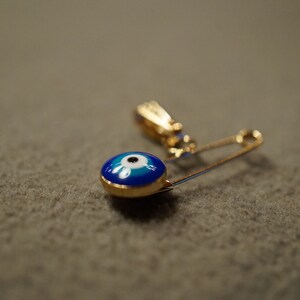 May include: A gold safety pin with a blue and white evil eye charm. The charm is round and has a white pupil in the center.
