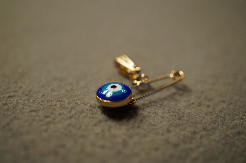 May include: A gold safety pin with a blue and white evil eye charm. The charm is round and has a white pupil in the center.