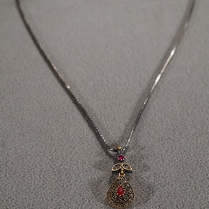May include: A silver chain necklace with a gold pendant featuring a red gemstone surrounded by smaller stones. The pendant is shaped like a teardrop with a small leaf design at the top.