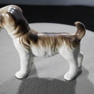 May include: A white porcelain figurine of a brown and white dog. The dog is standing on all four legs with its tail curled up. The figurine is in a side profile view.