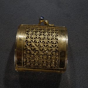 May include: A gold-colored metal trinket box with a lattice design. The box has a hinged lid and a small blue jewel on the top.