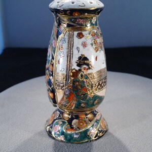 May include: A ceramic bottle stopper with a gold, green, blue and white floral design. The bottle stopper features a woman in a traditional Japanese kimono.