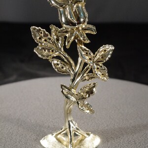 May include: A gold-toned metal rose with a detailed stem and leaves. The rose is on a small, round base.