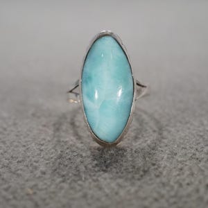 May include: A silver ring featuring a large, oval-shaped, light blue gemstone. The gemstone is set in a silver band and has a smooth, polished surface. The ring is displayed on a gray surface.