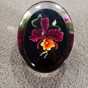K&ouml;nnte beinhalten: Ein ovaler, durchsichtiger Glascabochon mit schwarzem Hintergrund und einer handbemalten pinkfarbenen und orangefarbenen Orchideenbl&uuml;te.