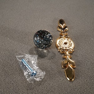 May include: A gold-toned cabinet knob with a decorative floral design and a clear crystal knob with a screw.
