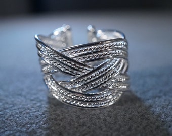 May include: A silver ring with a braided design. The ring is made of multiple strands of silver that are woven together to create a complex pattern.