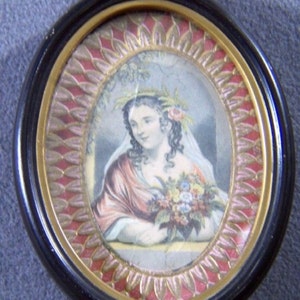 May include: An oval portrait miniature of a woman wearing a white veil and a wreath of flowers in her hair. She is holding a bouquet of flowers and is looking to the side. The portrait is framed in a black and gold oval frame with a red and gold patterned border.