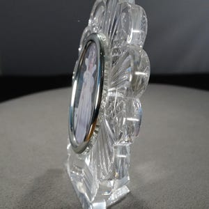 May include: A clear crystal picture frame with a silver-toned oval frame. The frame is shaped like a flower with a stand.