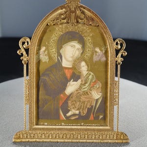 May include: A gold-toned framed religious icon depicting the Virgin Mary holding the Christ Child. The image features a gold background, with angels and text at the bottom. The frame has ornate filigree detailing and a decorative top.