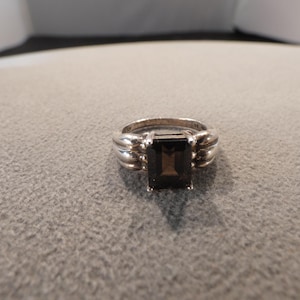 May include: A silver ring with a rectangular, dark brown gemstone. The ring has a ribbed design on either side of the gemstone, and the band appears to be smooth. The ring is set against a neutral gray background.