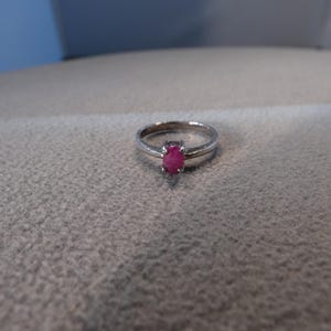 May include: A silver-toned metal ring featuring a small, oval-shaped, vibrant pink gemstone set in a prong setting. The band is smooth and polished, with a simple design that highlights the central pink stone as the primary focal point.