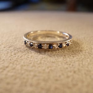 May include: A silver ring with a band of alternating blue and clear gemstones. The ring is set on a neutral-colored surface, showcasing the sparkle of the stones. A classic piece of jewelry.