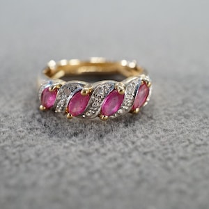 May include: A gold-toned ring with a series of alternating oval-shaped, bright pink gemstones and small, clear accent stones. The ring is set on a gray surface.