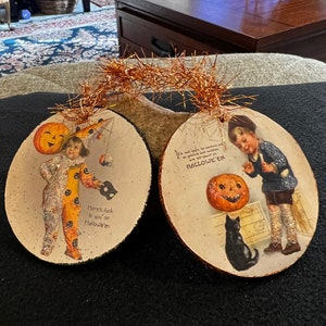 May include: Two round, wooden Halloween ornaments with glitter. One features a child in a jester costume holding a mask with the text "Here's luck to you on Hallowe'en". The other features a child holding a jack-o-lantern with a black cat and the text "It's not safe to venture out, as goblins and witches are all about HALLOWEEN."