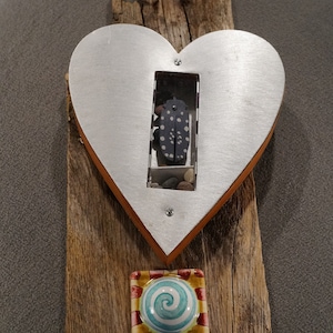 May include: A heart-shaped metal frame with a small window displaying a black and white spotted bug and small stones. The frame is attached to a piece of weathered wood with a colorful ceramic tile featuring a blue spiral design.
