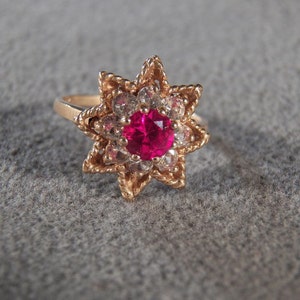 May include: A gold ring with a red gemstone surrounded by smaller white gemstones, set in a star-shaped design.