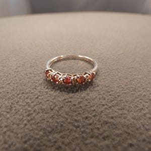 May include: A silver-toned band ring featuring a row of five round, vibrant orange-red gemstones set in individual prongs. The metal band is smooth and polished, showcasing a consistent circular shape with the stones arranged across the front arc.