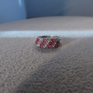 May include: A silver-toned ring featuring three horizontal rows of alternating round-cut pink and clear stones. The band has a polished metallic finish and the stones are set closely together in a multi-row cluster design across the front.