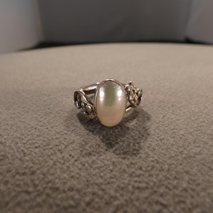 May include: A silver-toned ring featuring a central oval-shaped white pearl-like cabochon. The band is decorated with small metal floral motifs on both sides of the setting, creating a detailed and textured appearance around the central stone.