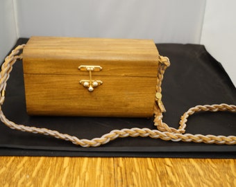 May include: A wooden box purse with a braided strap. The box is brown and has a gold latch. The strap is a light brown and white braid.