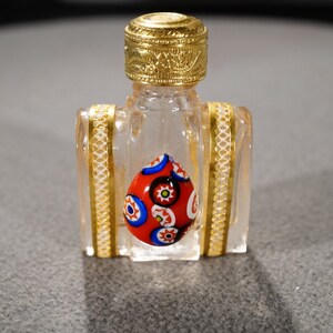 May include: A small, clear glass bottle with a gold top and a red, blue, and white glass bead inside. The bottle has a gold trim around the edges.