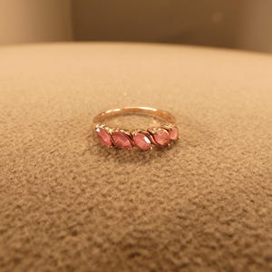 May include: A rose gold ring featuring five oval-cut pink gemstones. The gemstones are set in a row across the top of the ring. The ring is displayed on a neutral-colored surface.