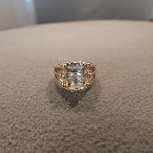 May include: A gold-toned ring featuring three horizontally aligned, marquise-cut, pale blue gemstones. The band is constructed with multiple thin, parallel gold-colored metal wires that converge at the sides to support the central stone setting.