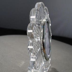 May include: A clear crystal picture frame with a scalloped edge and a silver-toned metal frame. The frame is designed to hold a small oval-shaped photo.