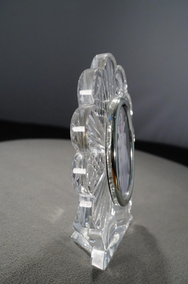 May include: A clear crystal picture frame with a scalloped edge and a silver-toned metal frame. The frame is designed to hold a small oval-shaped photo.