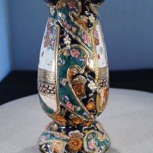 May include: A decorative ceramic vase with a floral pattern in shades of green, gold, pink, and white. The vase has a wide base and a narrow neck.