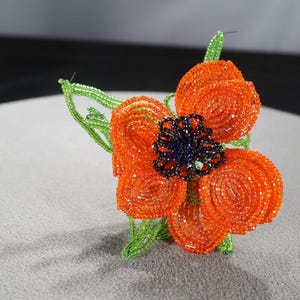 May include: A beaded flower with orange petals, a dark blue center, and green stems and leaves. The flower is made of small, round beads and is likely a decorative item or accessory.