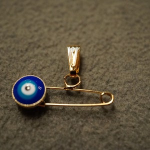May include: A gold safety pin with a blue and white evil eye charm attached to the top.