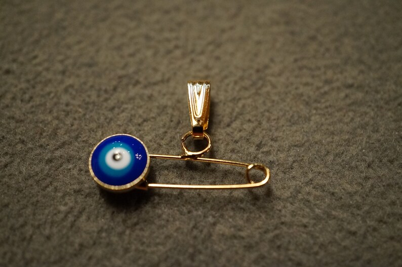 May include: A gold safety pin with a blue and white evil eye charm attached to the top.
