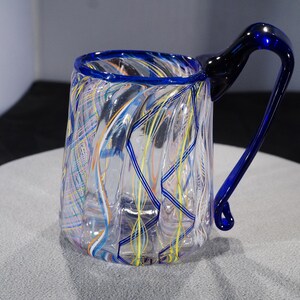 May include: A clear glass mug with a blue rim and handle. The mug has a swirling pattern of colorful glass threads throughout.