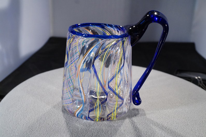 May include: A clear glass mug with a blue rim and handle. The mug has a swirling pattern of colorful glass threads throughout.