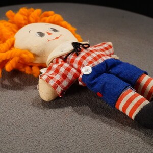 May include: A rag doll with orange yarn hair, a red and white checkered shirt, blue pants, and red and white striped socks. The doll has a painted face with black eyes and a red mouth.