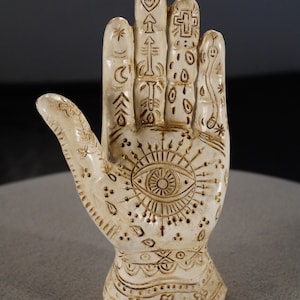 May include: A white ceramic hand sculpture with intricate brown designs, including an eye with radiating lines, a heart, a cross, and other symbols.