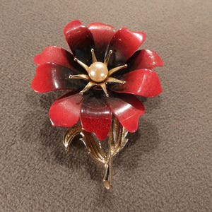 May include: A floral brooch featuring a red and black flower with a gold-tone center and stem. The flower has a pearl-like bead in the center. The brooch is a vintage accessory.