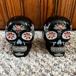 May include: Two black sugar skull salt and pepper shakers with colourful floral designs. The shakers are sitting on a grey carpet.