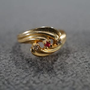 May include: A gold-toned ring with a swirling design. The ring features a small, round, red gemstone in the center, flanked by two smaller clear gemstones. The band has a textured, layered appearance.