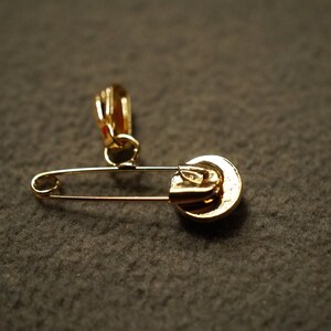 May include: A gold safety pin with a decorative charm attached to the top. The charm is a round, gold-colored disc with a raised design.