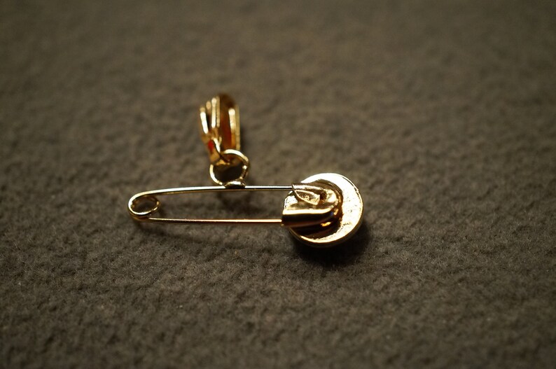 May include: A gold safety pin with a decorative charm attached to the top. The charm is a round, gold-colored disc with a raised design.