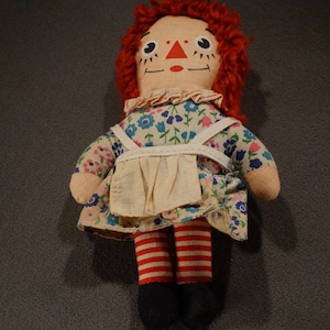 May include: A vintage rag doll with red yarn hair, a floral dress, a white apron, and red and white striped stockings. The doll has a painted face with black eyes, a red nose, and a smiling mouth.