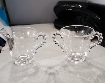 May include: Two clear glass creamer and sugar bowls with beaded handles. The bowls are empty and sitting on a gray surface.
