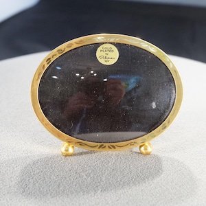 May include: Gold-plated oval picture frame with a dark interior. The frame has a decorative border and rests on three small, rounded feet. The text "GOLD PLATED by Pilken" is visible on a small sticker.