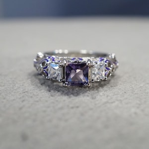 May include: A silver ring with a purple square-shaped gemstone surrounded by two smaller clear square-shaped gemstones. The band of the ring is decorated with a blue and white floral design.