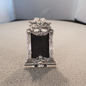 May include: A small, ornate silver-colored picture frame with a black velvet backing. The frame features intricate detailing, including floral and scrollwork designs, and a decorative top with two cherub figures. The frame is rectangular and stands upright.
