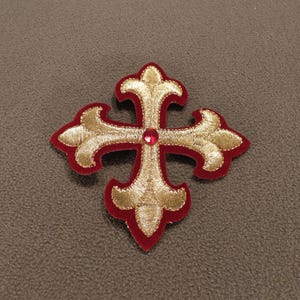 May include: A cross-shaped patch featuring gold metallic embroidery on a deep red velvet base. The design includes a fleur-de-lis style at each of the four ends and a single, small, round, bright red gemstone set at the center of the cross.