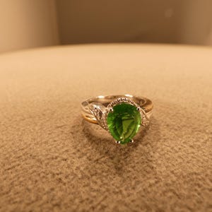May include: A silver and gold ring with a large, oval-cut, bright green gemstone as the centerpiece. The ring has a halo of small, clear stones around the main gem and additional accent stones on the band.